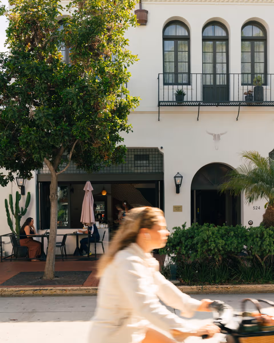 Woman cycling past Spanish colonial style building with outdoor cafe, white stucco walls, arched windows, and palm trees at