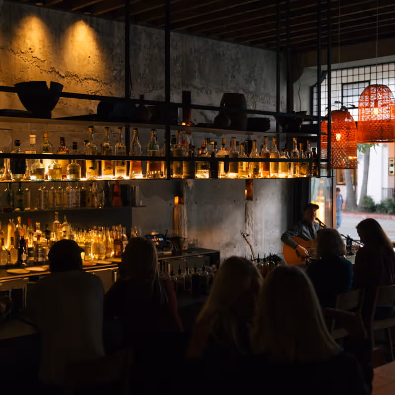 Intimate bar interior with live acoustic guitar performance, patrons seated at counter, backlit liquor shelves and woven