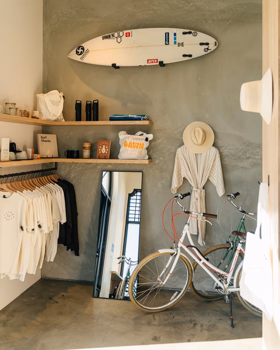 Coastal surf shop interior with white surfboard mounted on concrete wall, clothing rack with t-shirts, vintage bicycles, and