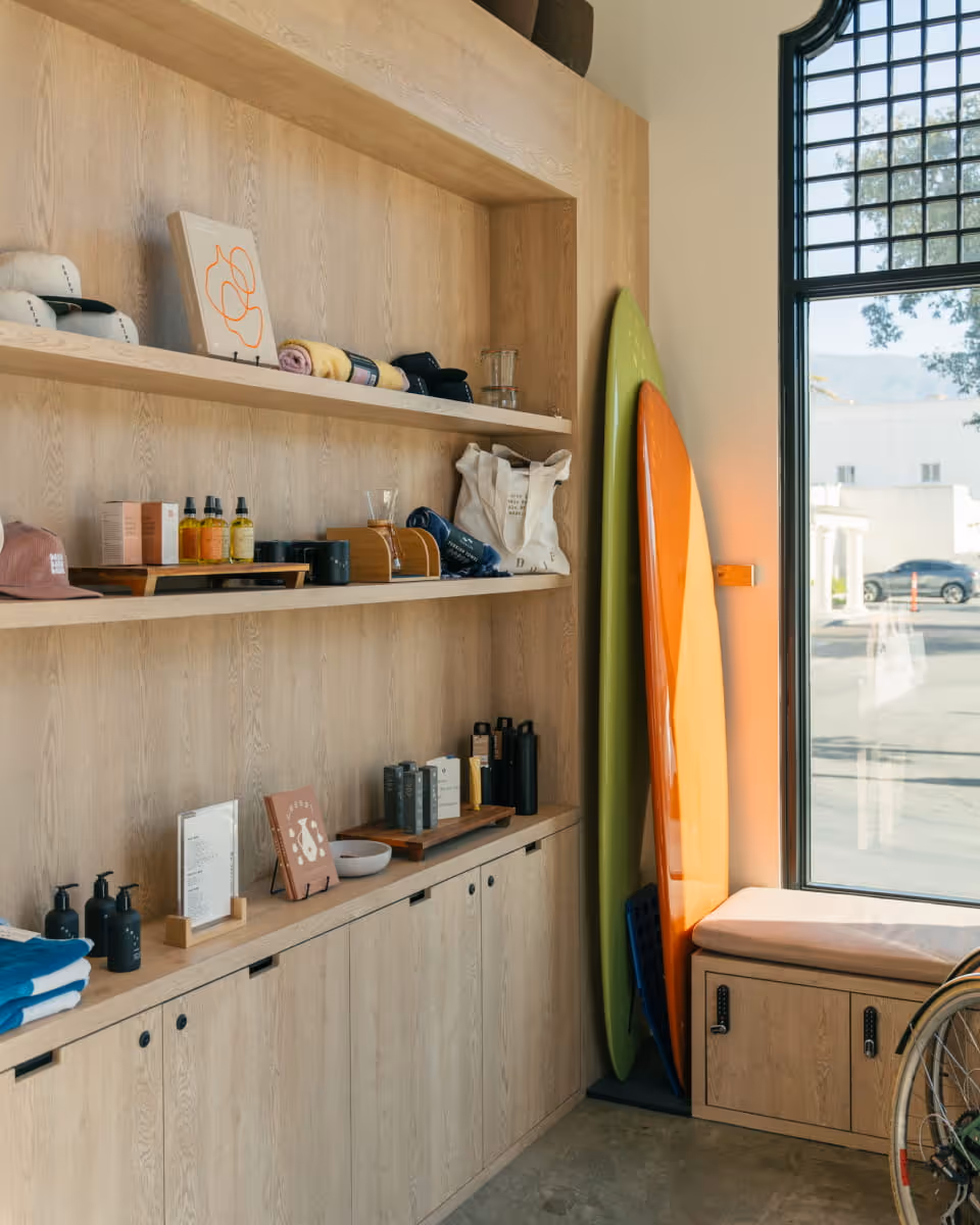 Coastal retail shop display with light oak shelving featuring surfboards, skincare products, beach towels, and lifestyle