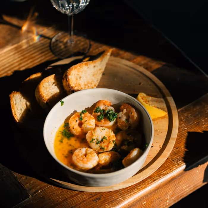 Gambas al ajillo garlic butter shrimp in white bowl with toasted bread slices and lemon wedge on wooden serving board