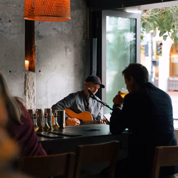 Live acoustic music performance at intimate bar venue with guitarist singing into microphone while patrons enjoy drinks