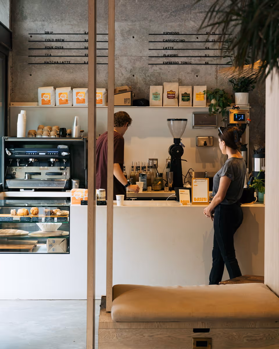 Modern Dawn cafe interior with concrete walls, espresso machine, pastry display case, and menu board listing coffee drinks