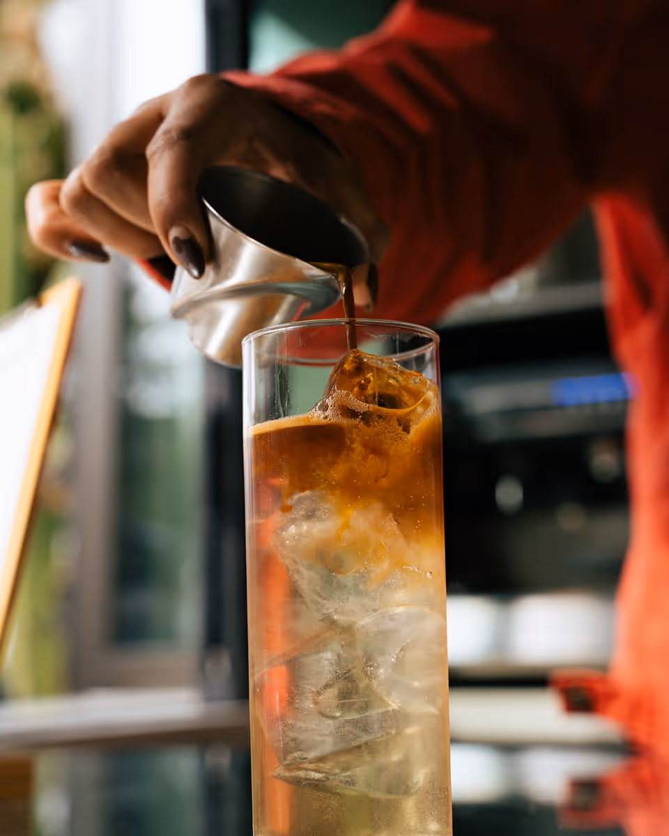 Barista pouring espresso shot from metal pitcher into tall glass with ice, creating layered iced coffee drink