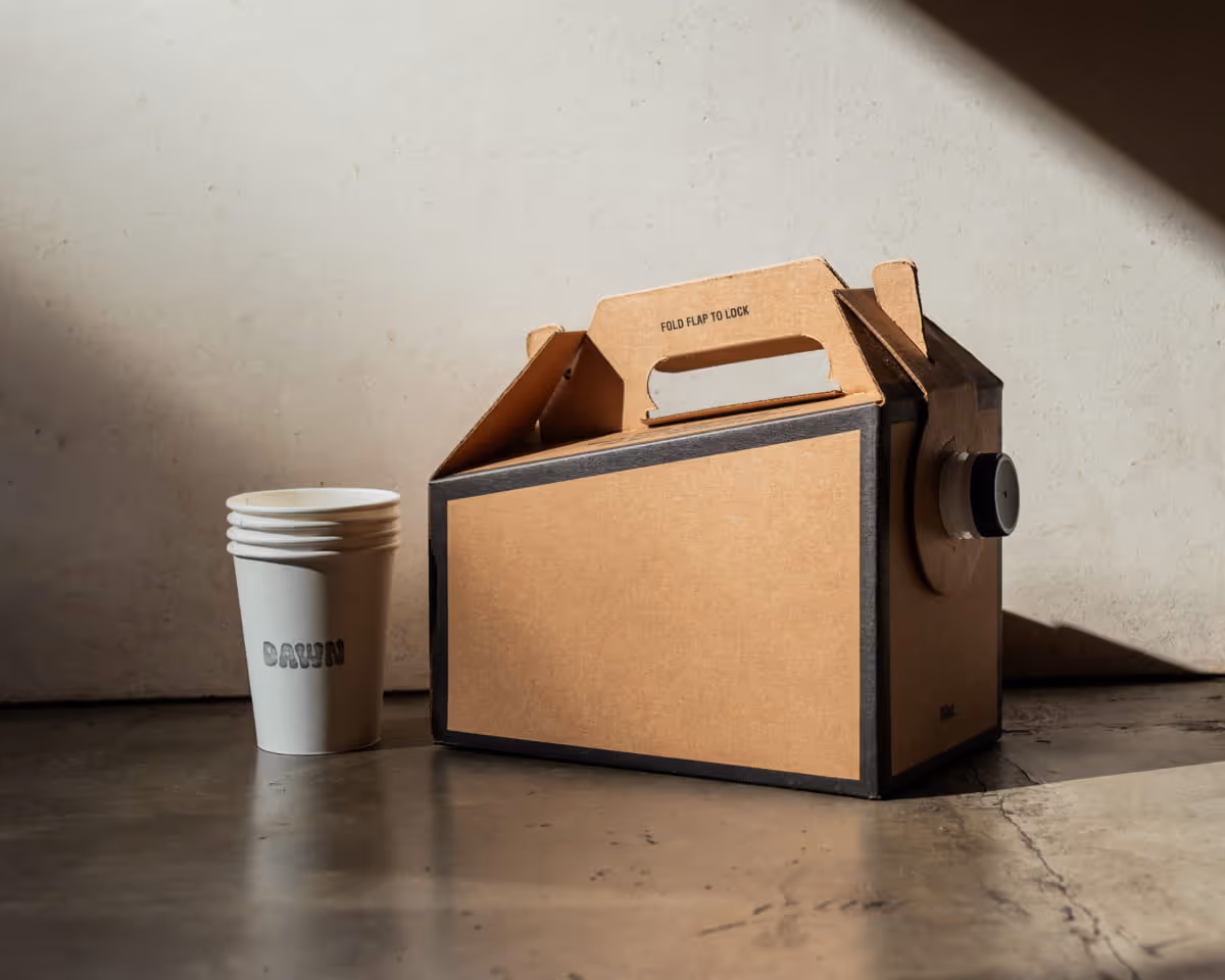 Dawn coffee box carrier with fold flap to lock handle and stacked white paper cups on concrete surface with natural light