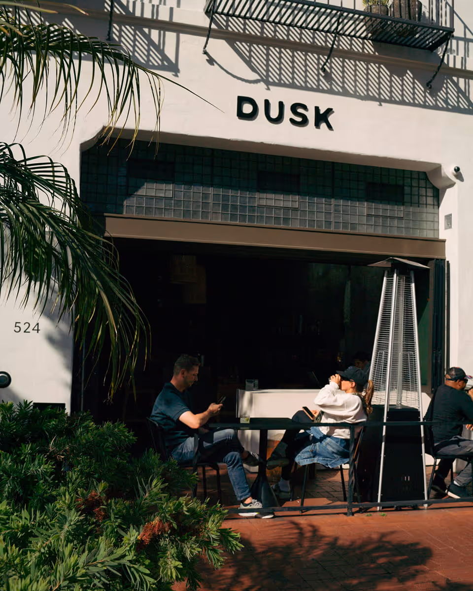 DUSK restaurant exterior at 524 with outdoor patio seating, palm trees, and customers enjoying sunny California day