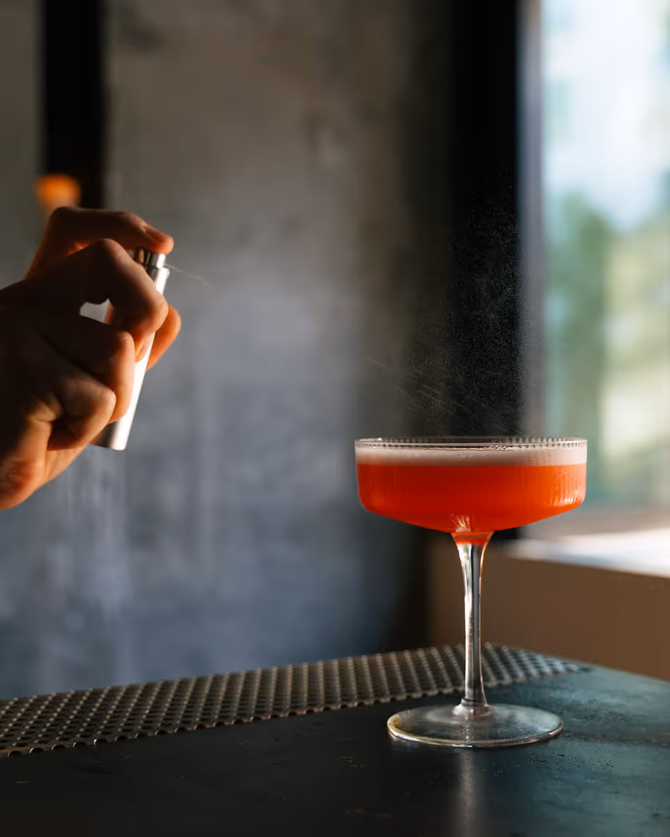 Bartender spraying aromatic mist over elegant red cocktail in coupe glass on bar counter with soft natural light