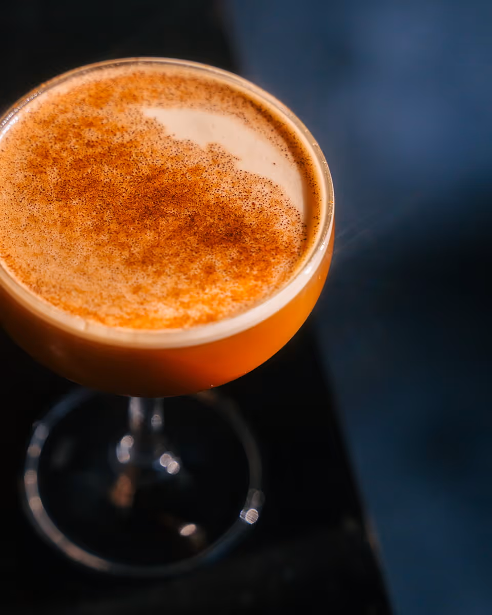 Elegant cocktail in coupe glass with creamy foam top dusted with cinnamon spice, orange-hued drink on dark bar surface