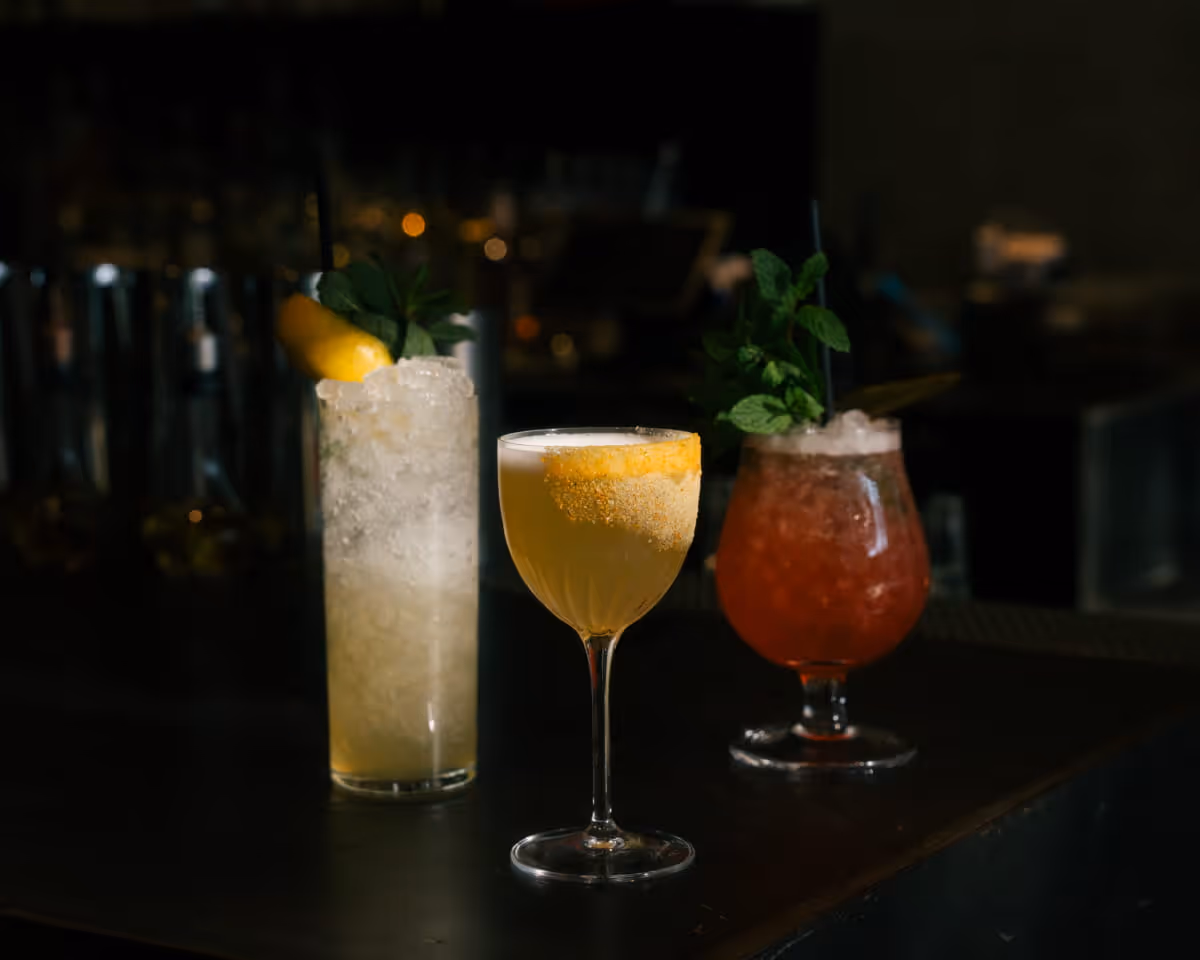 Three craft cocktails on bar counter featuring a highball with lemon, golden coupe drink, and red frozen cocktail with mint
