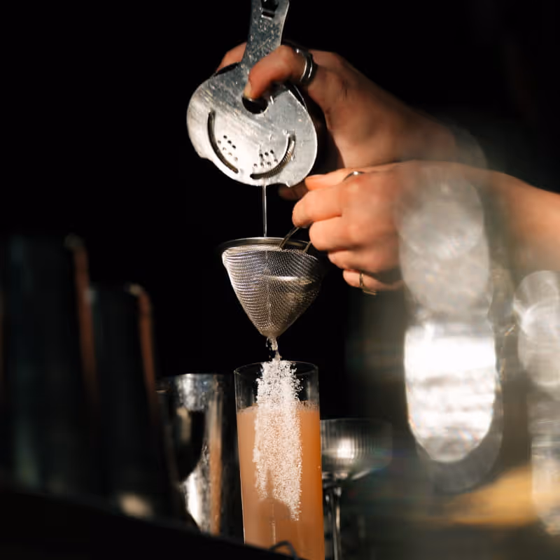 Bartender double straining a pink cocktail through a fine mesh strainer into a tall glass at a dimly lit bar