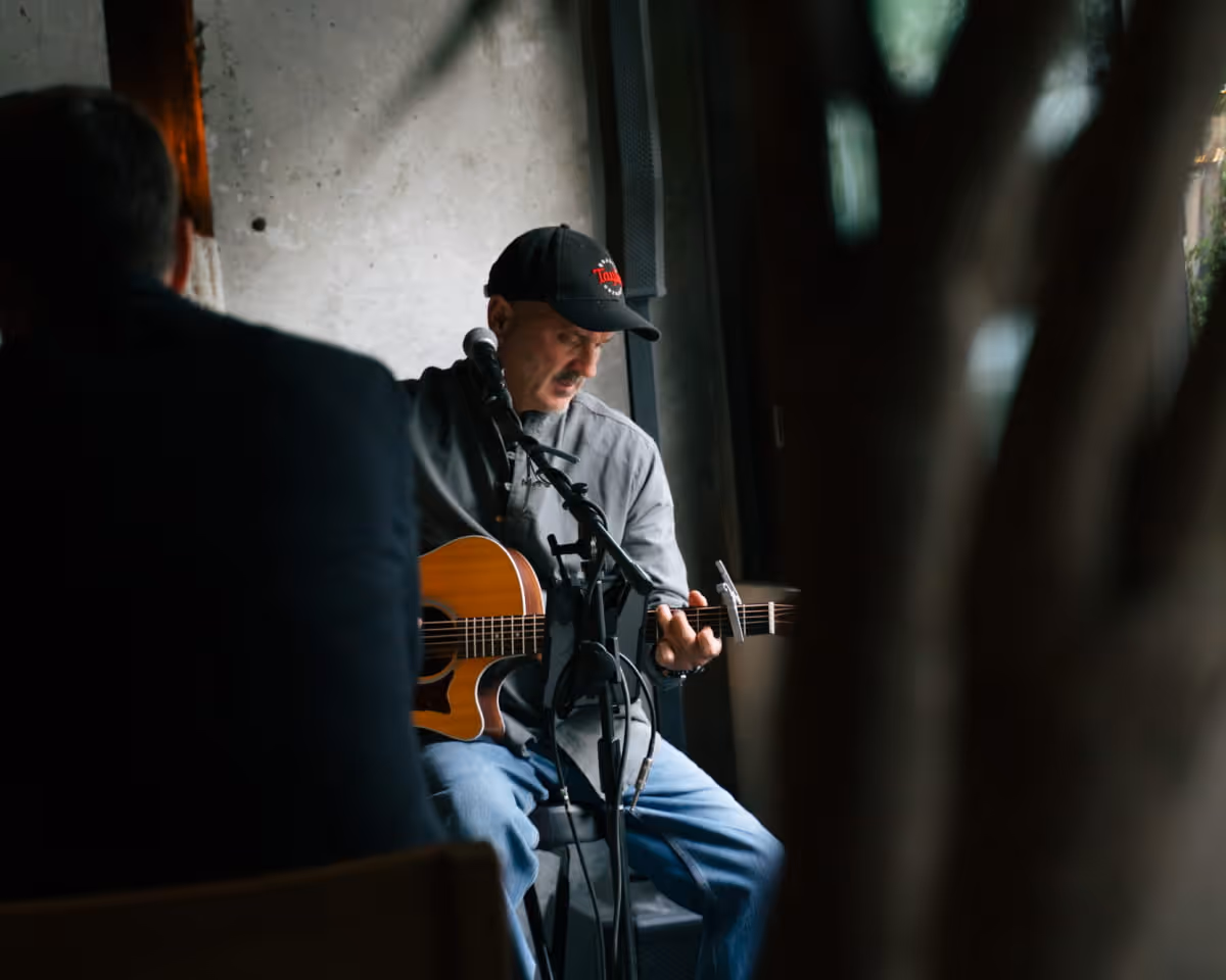 Acoustic guitarist wearing Taylor cap performing live music at intimate bar venue with microphone setup and natural lighting