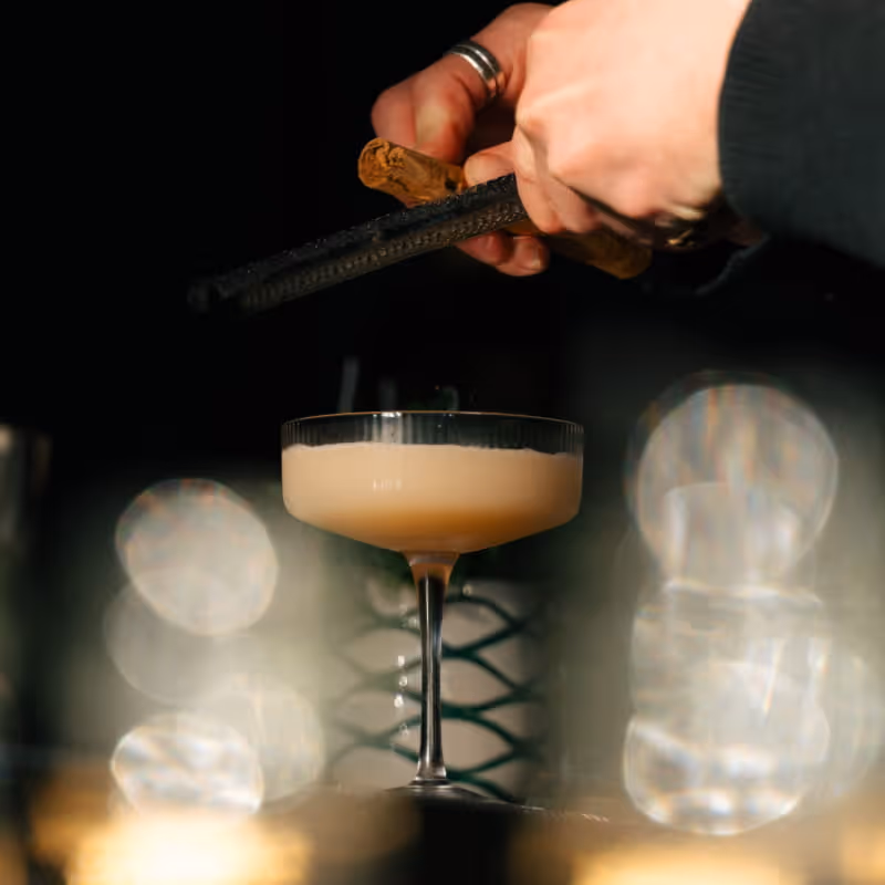 Bartender grating cinnamon or nutmeg over a creamy coupe cocktail with elegant bokeh lighting in upscale bar setting