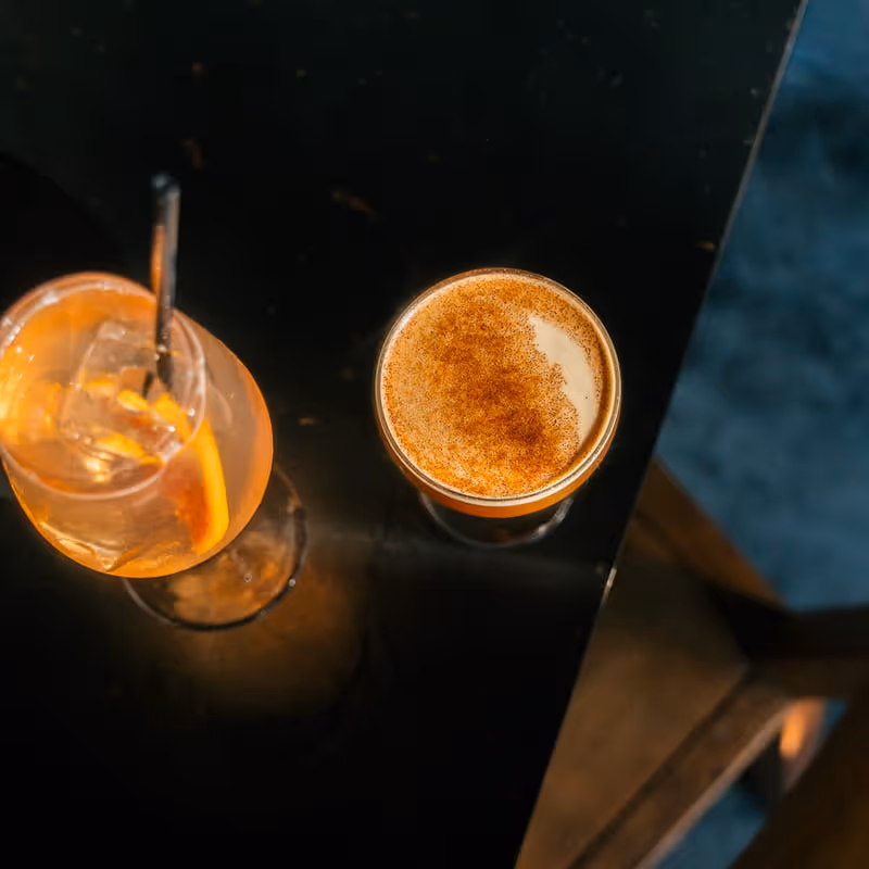 Two craft cocktails on dark bar counter - orange spritz with ice and straw, whiskey sour with cinnamon foam topping