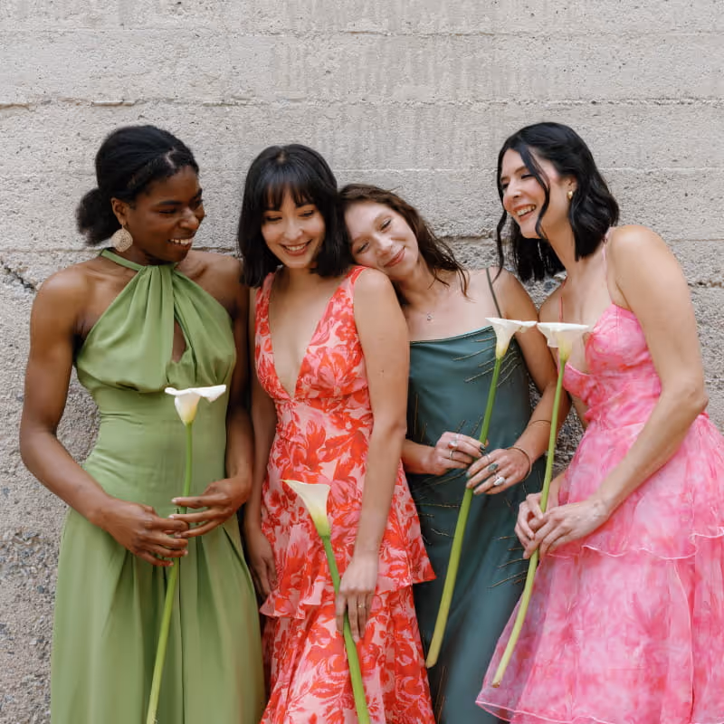 Four women in colorful spring dresses holding white calla lilies, wearing green, coral floral, emerald, and pink gowns