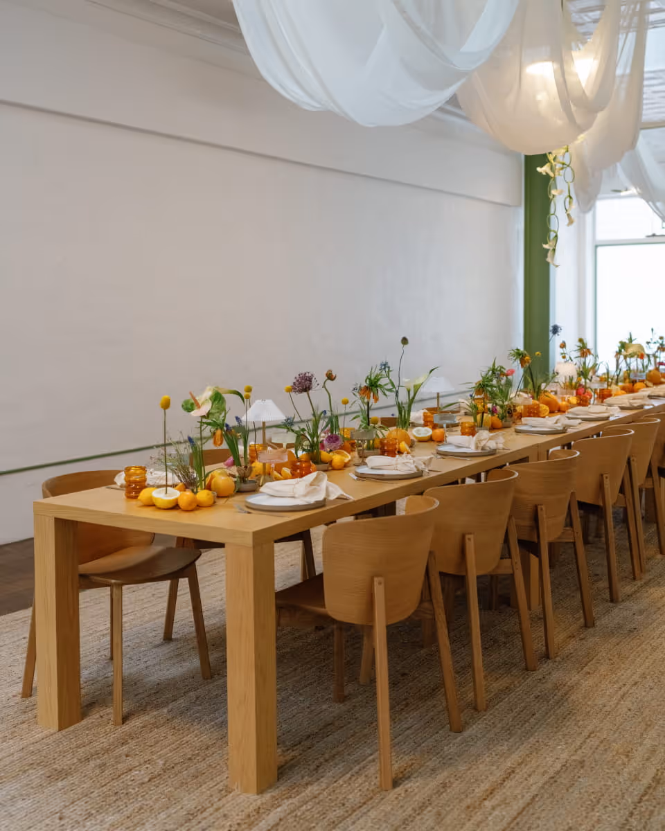 Elegant long wooden dining table with citrus centerpieces, wildflowers, amber glassware, and white fabric ceiling draping