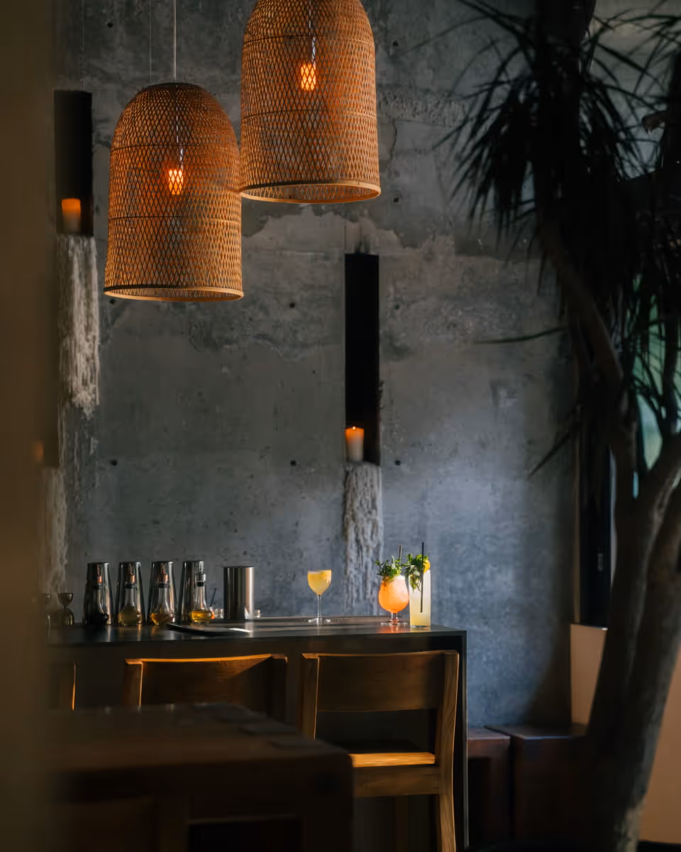 Moody cocktail bar interior with woven rattan pendant lights, concrete walls, colorful craft cocktails on dark wooden counter