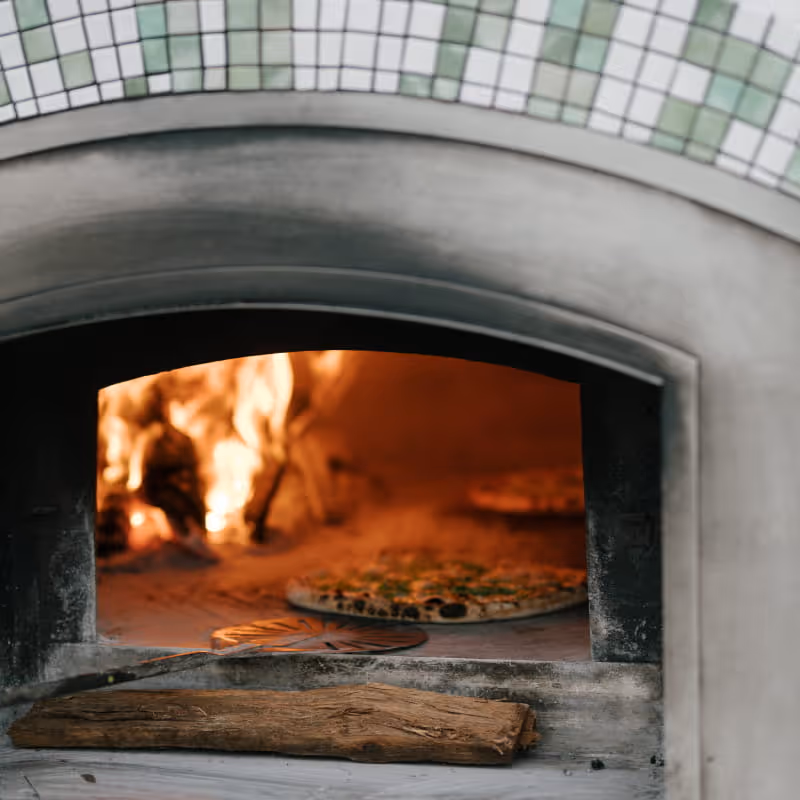 Wood-fired pizza oven with flames cooking fresh pizza, wooden peel and log in foreground, decorative tile arch