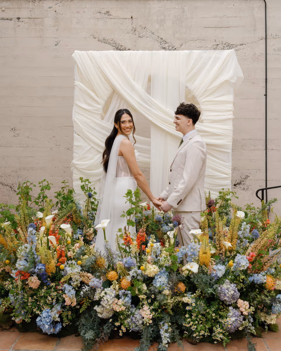 Bride and groom holding hands at ceremony altar surrounded by vibrant wildflower arrangement with white draped fabric