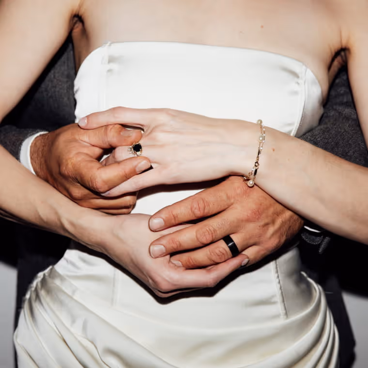 Intimate wedding couple embrace showing rings and jewelry, bride in white satin dress with groom's arms wrapped around