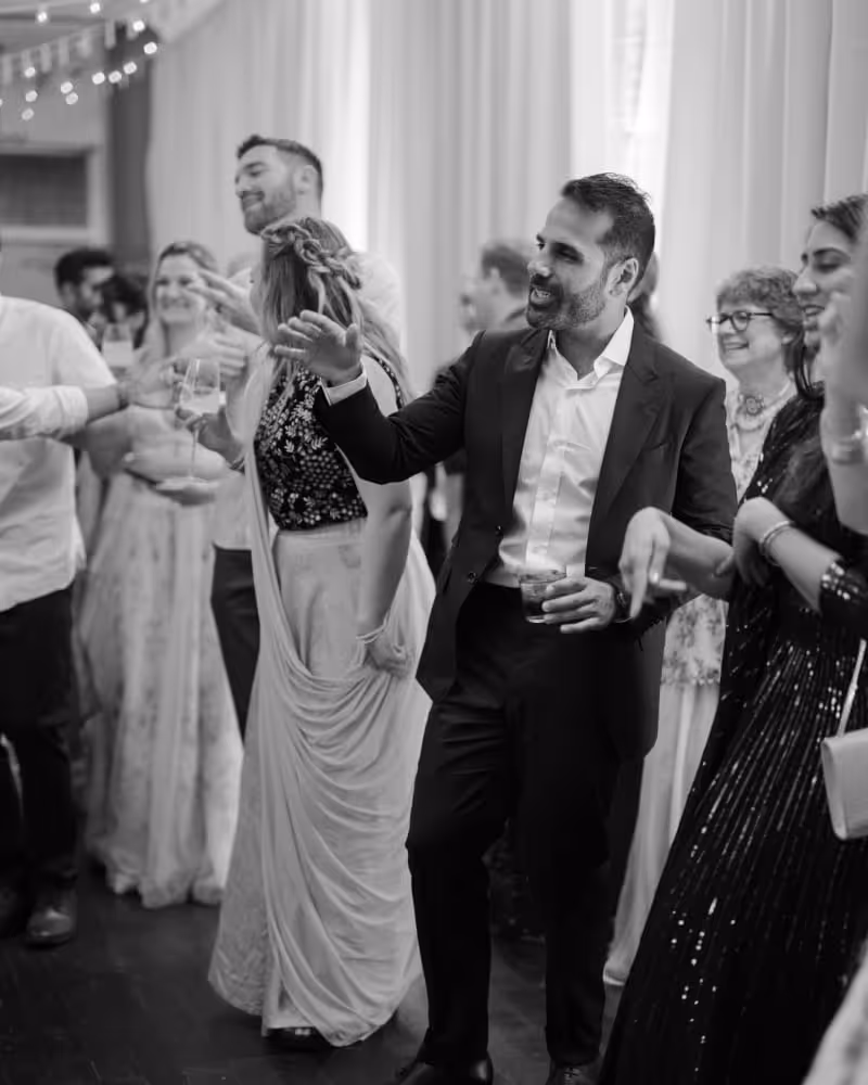 Wedding guests dancing and celebrating on dance floor, man in dark suit holding drink enjoying party atmosphere