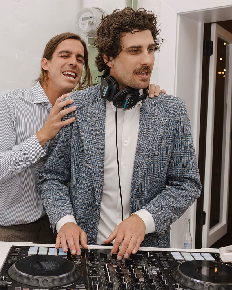 DJ in plaid blazer mixing music on turntables while friend laughs behind him at wedding reception entertainment