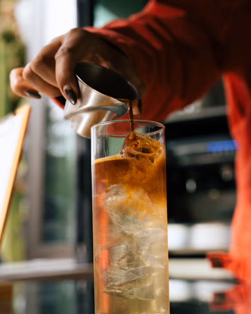 Barista pouring espresso shot over ice in tall glass, creating layered iced coffee drink at specialty coffee bar