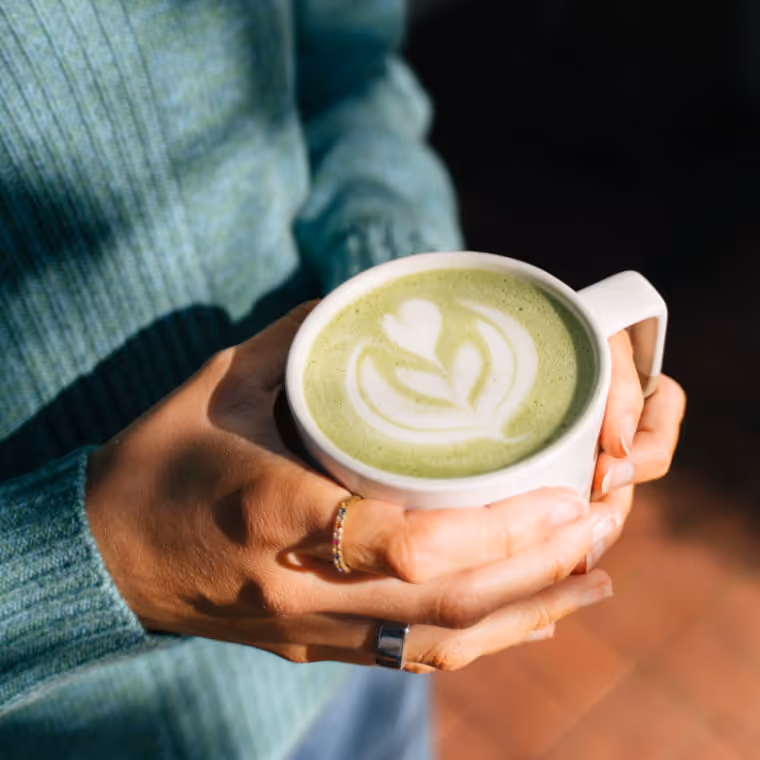 Hands holding white ceramic mug with matcha latte featuring delicate leaf latte art, cozy sage green sweater sleeves