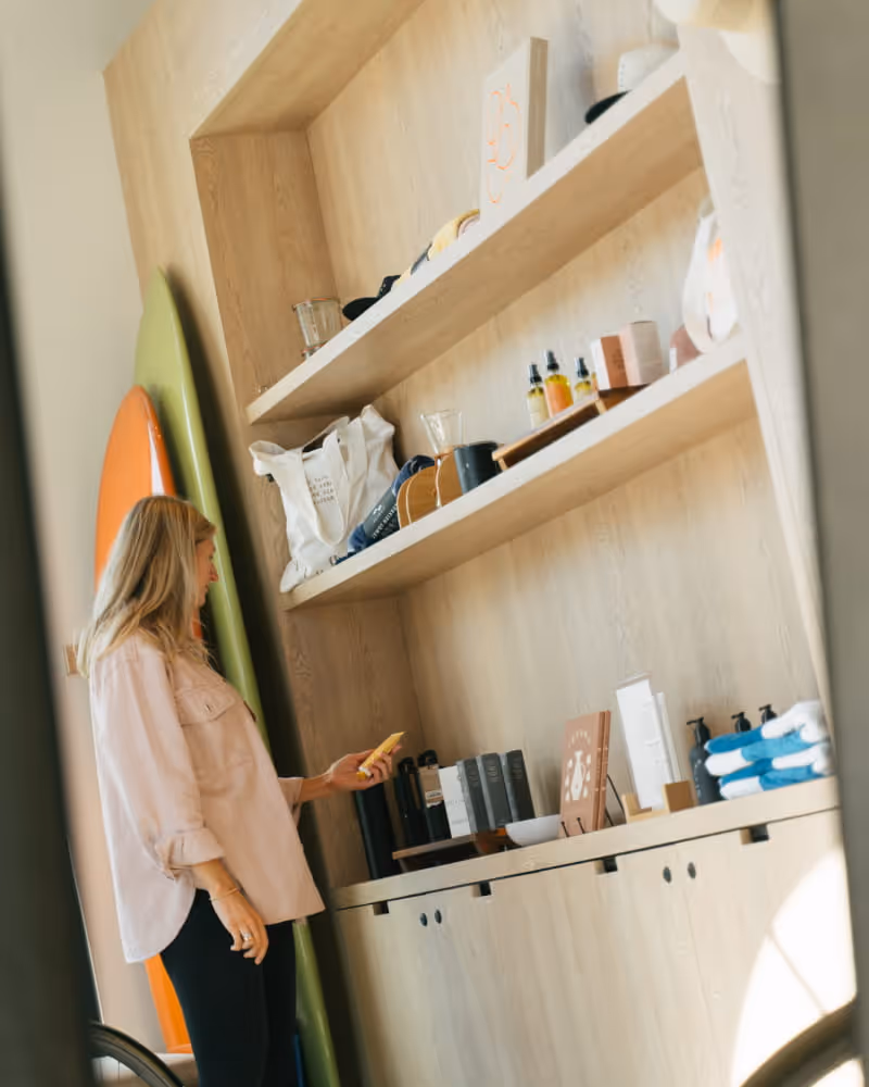 Woman browsing curated lifestyle products on wooden shelves in hotel retail shop with surfboards and skincare items displayed