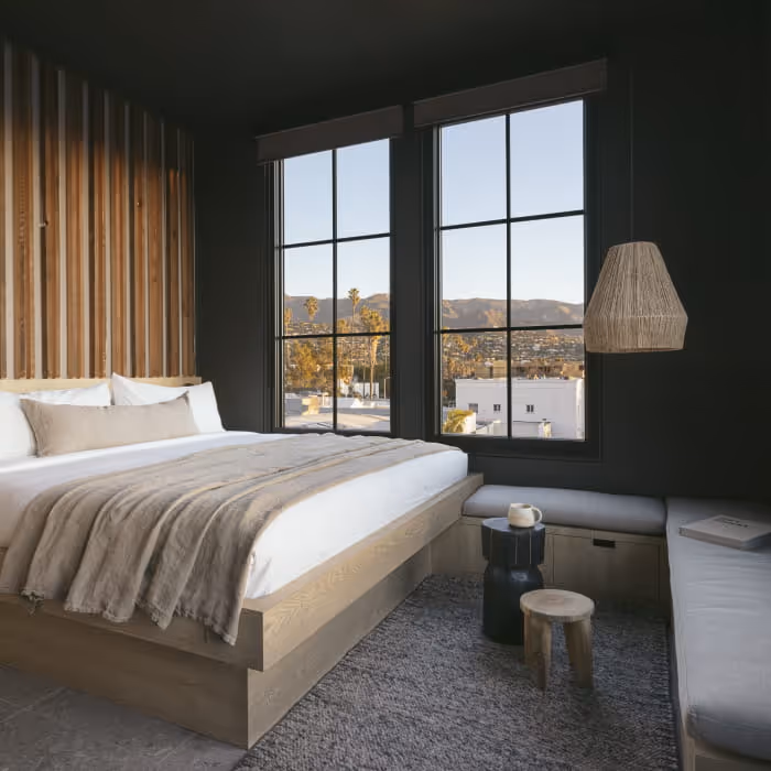 Modern boutique hotel bedroom with dark walls, wooden slat accent, platform bed with neutral linens, and mountain view