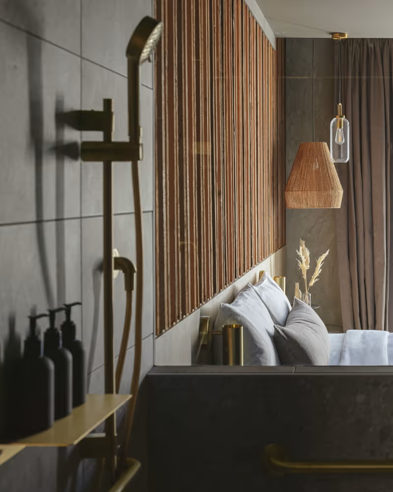 Luxury open-concept bathroom with dark stone soaking tub, brass fixtures, slatted wood accent wall, and view into bedroom