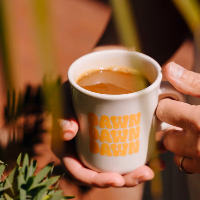 Hands holding a white ceramic coffee cup with "DAWN" repeated three times in yellow retro lettering, surrounded by green