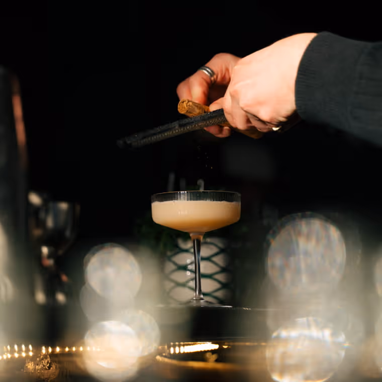 Bartender grating garnish over an elegant coupe cocktail with creamy foam top, atmospheric bokeh lights in dark bar setting