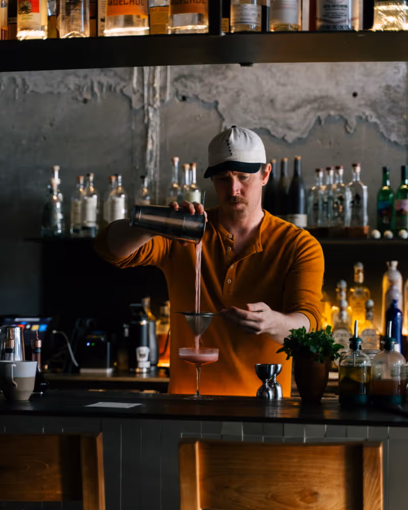 Bartender in orange polo shirt and cap pouring pink cocktail from shaker into coupe glass at craft bar with backlit bottles