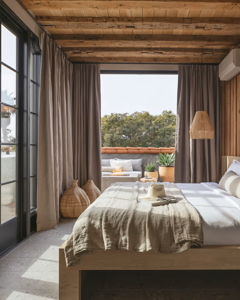 Rustic modern bedroom with wooden ceiling, linen bedding, woven vases, and outdoor terrace with fire pit and greenery