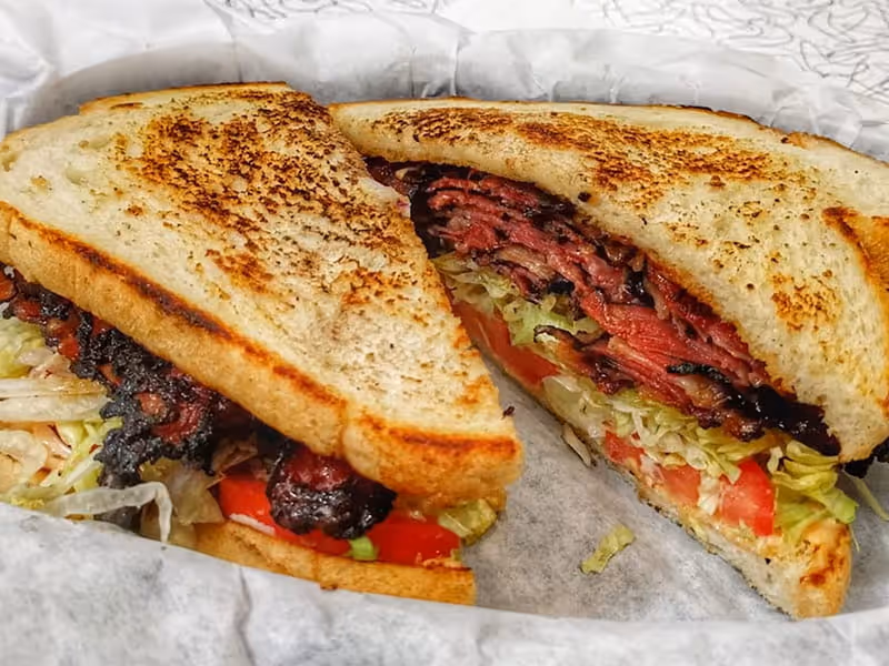 Toasted pastrami sandwich with shredded lettuce and tomatoes on grilled white bread served on deli paper