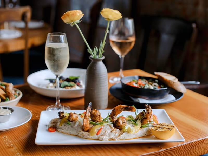 Grilled shrimp and grits platter with charred lemon on elegant restaurant table with wine glasses and yellow ranunculus