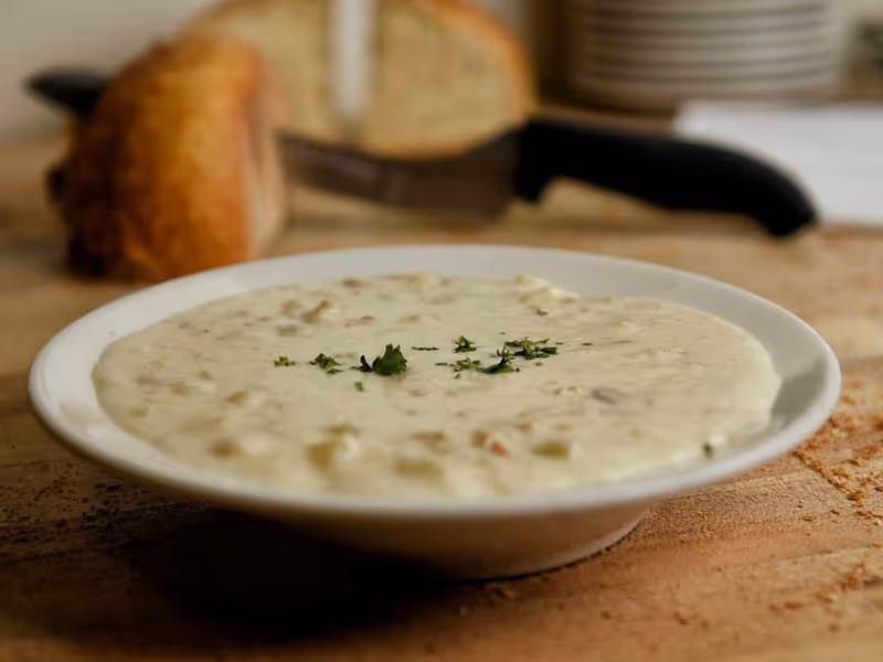 Creamy clam chowder soup garnished with fresh parsley in white bowl, served with rustic bread on wooden table