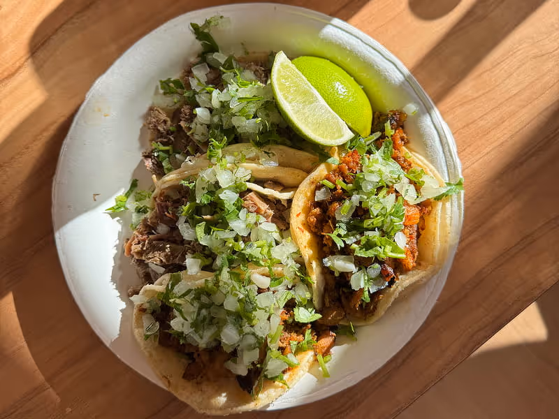 Three authentic Mexican street tacos with carnitas and al pastor, topped with cilantro, onions, and fresh lime wedges