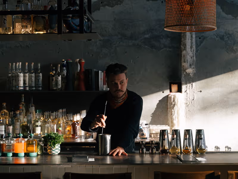 Bartender with mustache stirring cocktail in metal shaker at industrial-style craft cocktail bar with liquor bottles on