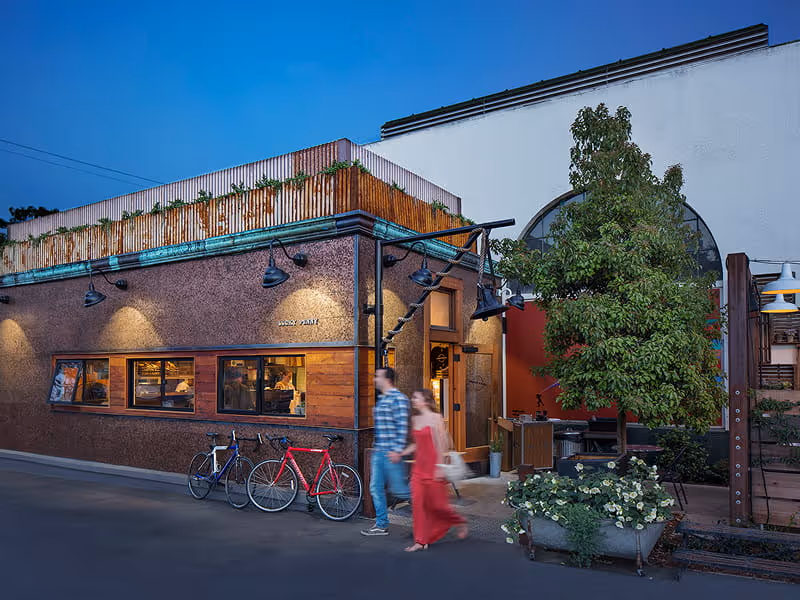 Trendy urban restaurant exterior at twilight with warm lighting, bicycles parked outside, and couple walking on sidewalk