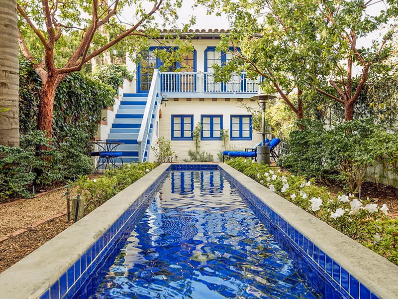 Mediterranean-style courtyard with vibrant blue lap pool, white stucco home with blue shutters, and lush garden landscaping