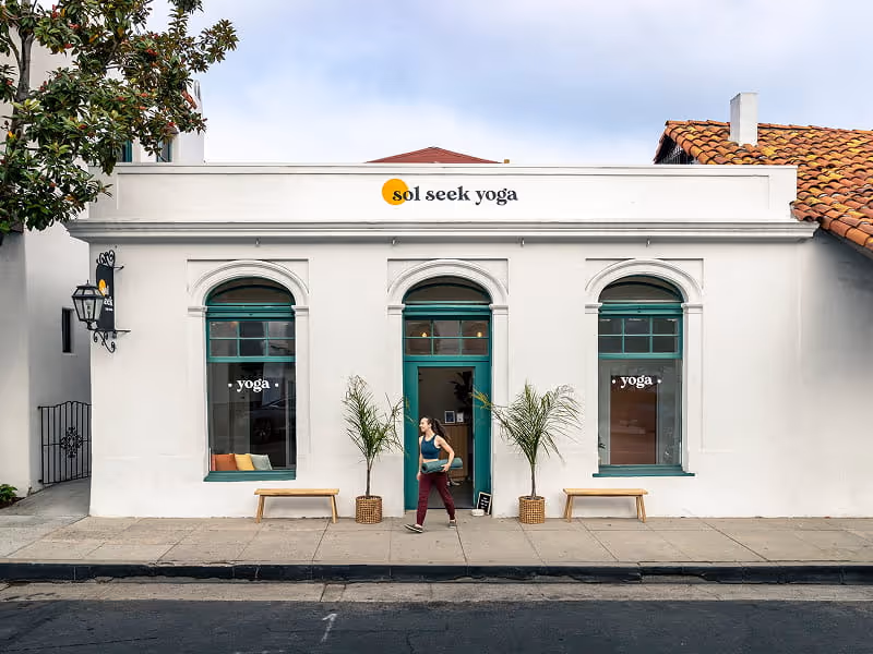 Sol Seek Yoga studio storefront with white Spanish-style architecture, teal trim, arched windows, and woman exiting with