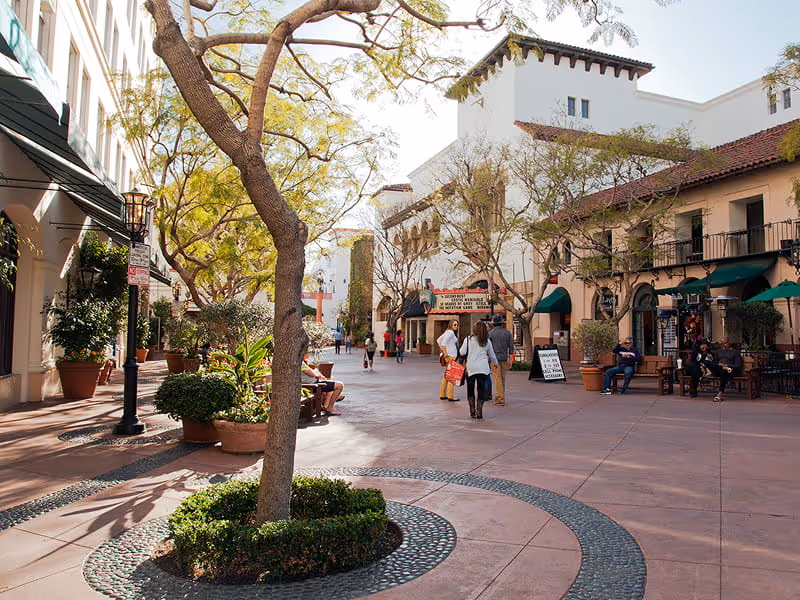 Pedestrians walking through Spanish-style outdoor shopping plaza with terracotta tiles, potted plants, and cafe seating in