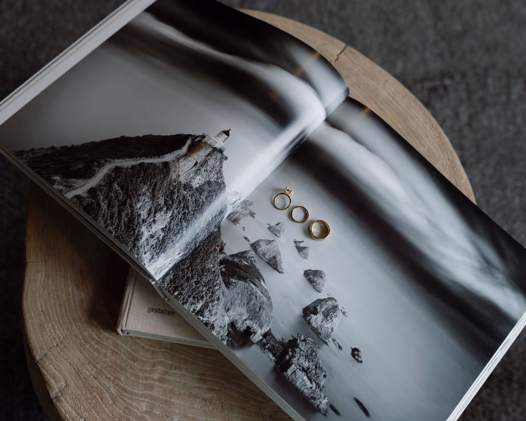 Three gold wedding rings placed on an open Gestalten photography book featuring a dramatic coastal cliff and lighthouse