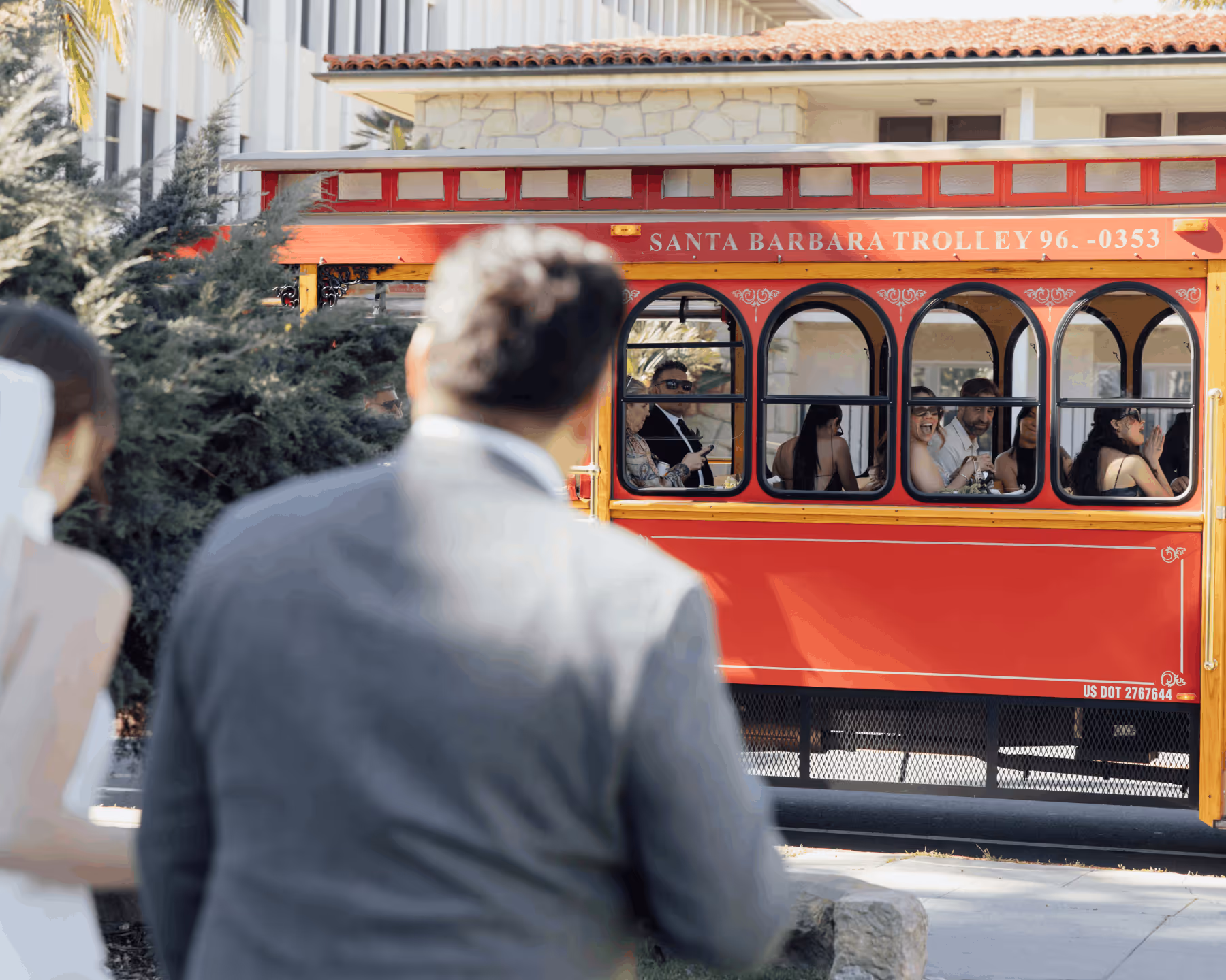 Wedding guests wave excitedly from a red Santa Barbara Trolley as the bride and groom watch from outside the vehicle.
