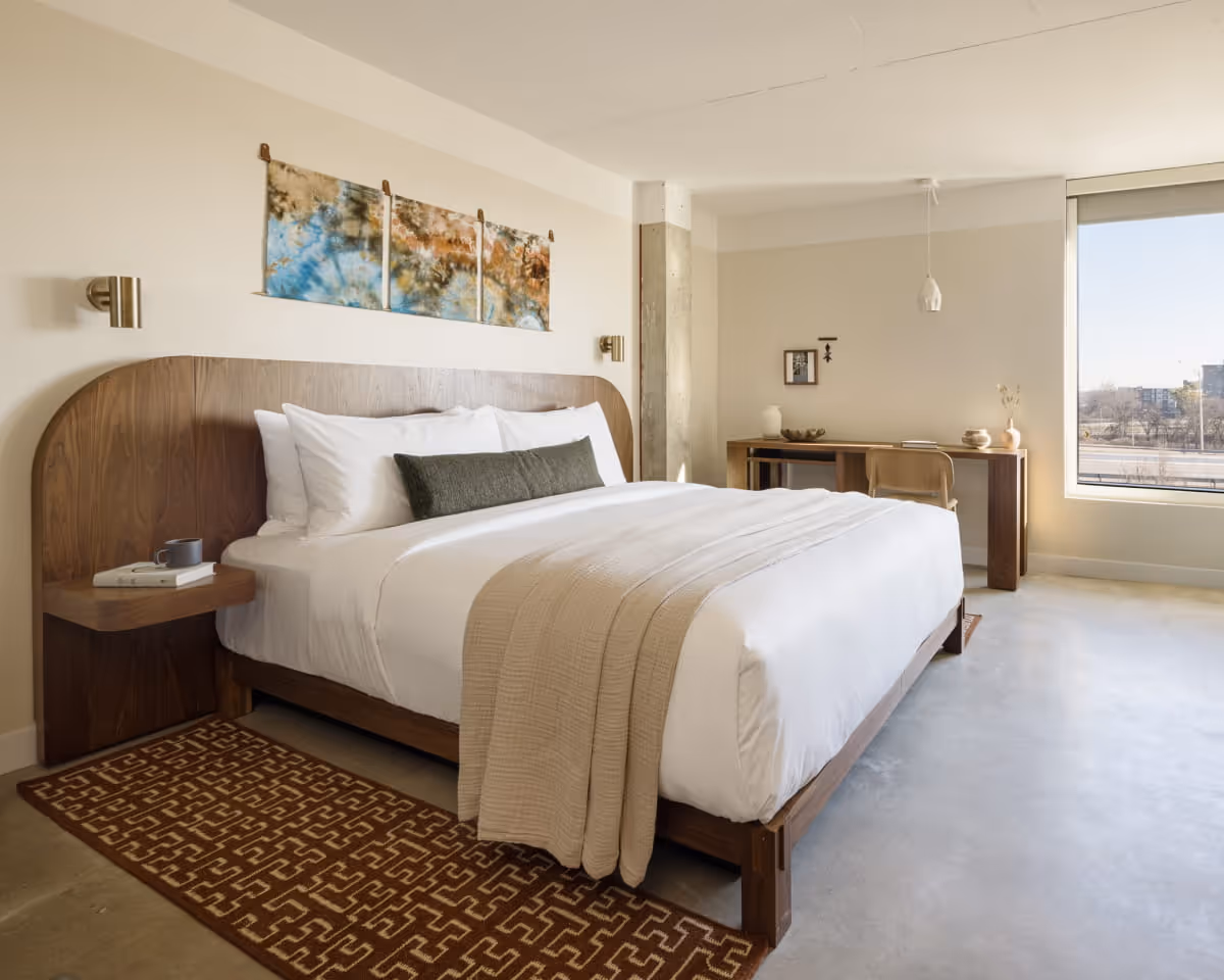 Modern hotel bedroom featuring a walnut wood bed frame with curved headboard, white bedding, geometric area rug, and large