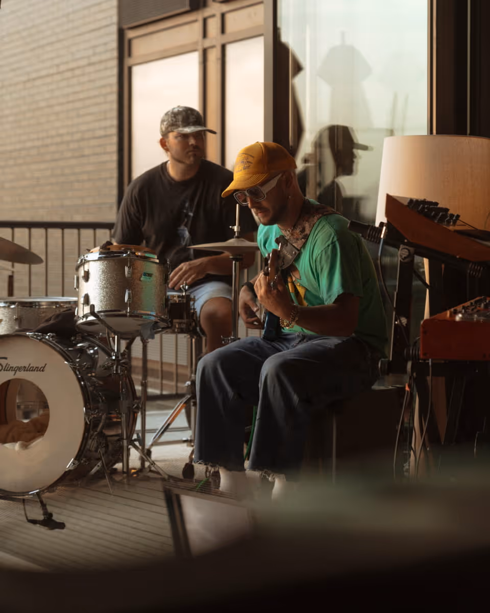 Two musicians jamming indoors, one playing guitar in a yellow cap and green shirt, another on a Slingerland drum kit.
