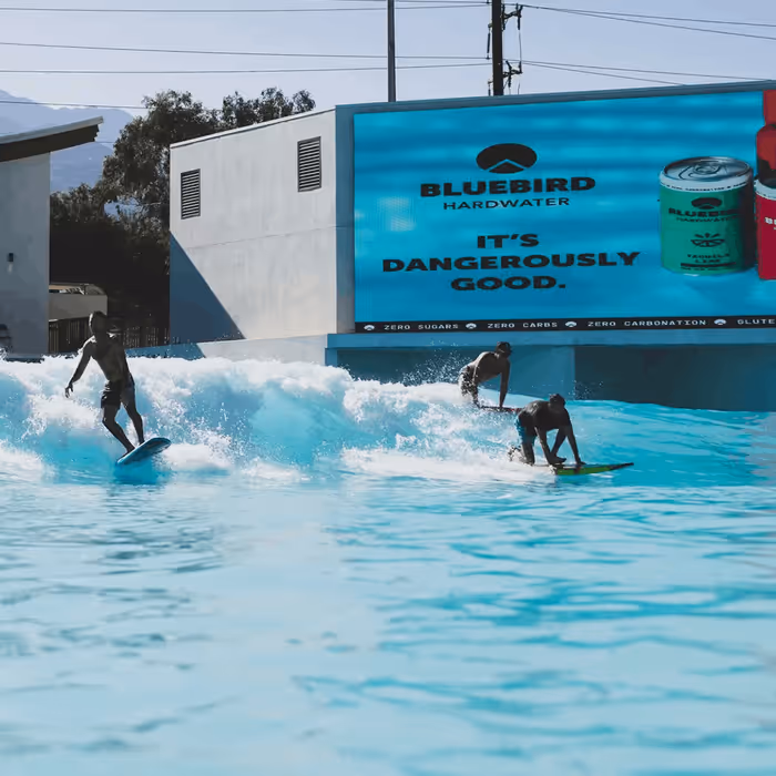 Surfers riding artificial waves at Palm Springs Surf Club with Bluebird Hardwater advertisement on large screen
