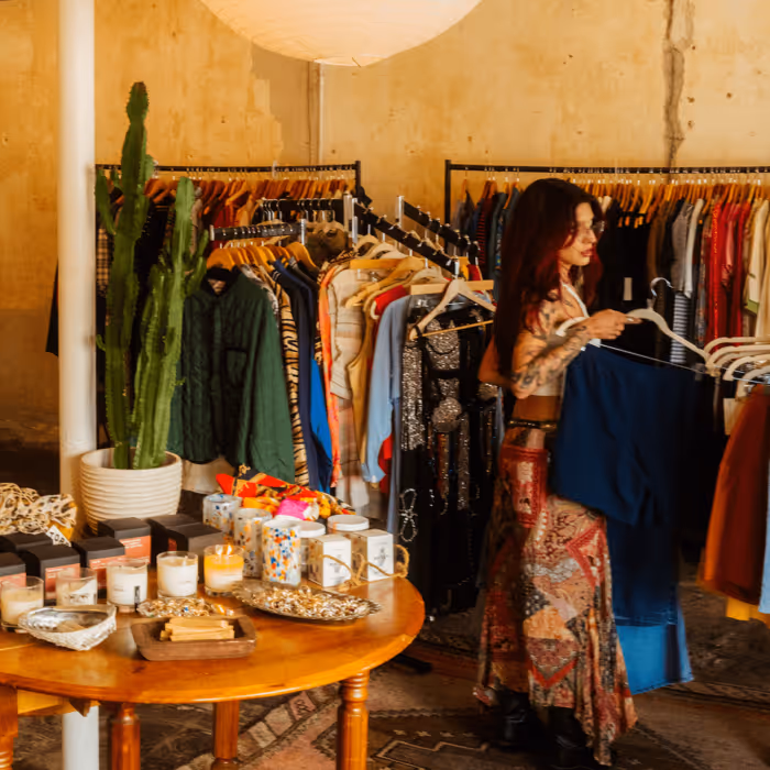 Woman in bohemian dress browses clothing racks in a vintage boutique with candles and accessories on display.