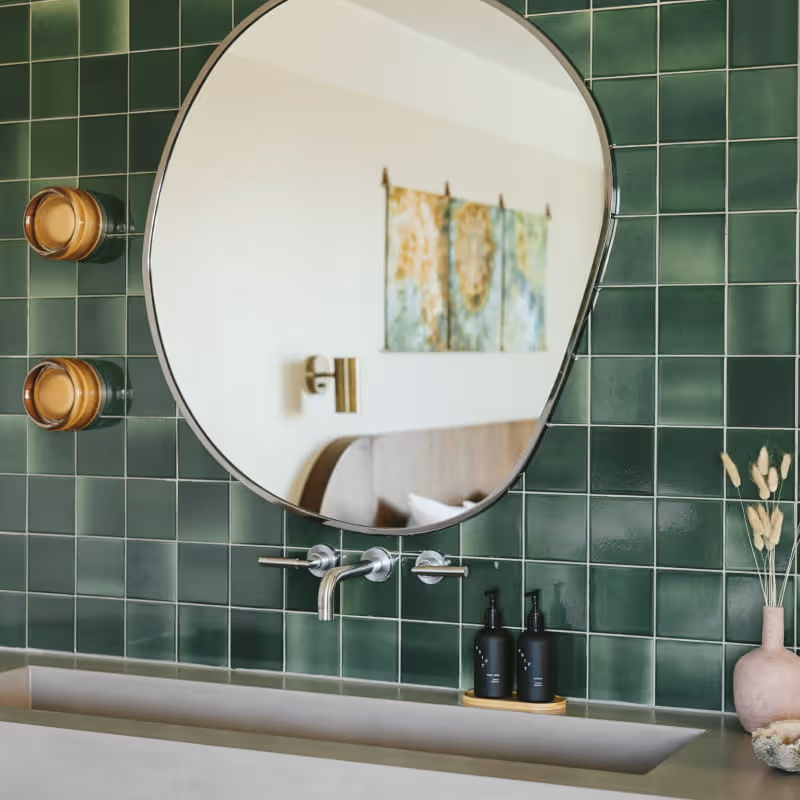 Bathroom vanity with deep green square tiles, asymmetrical round mirror reflecting bedroom, wall-mounted faucet, and amber